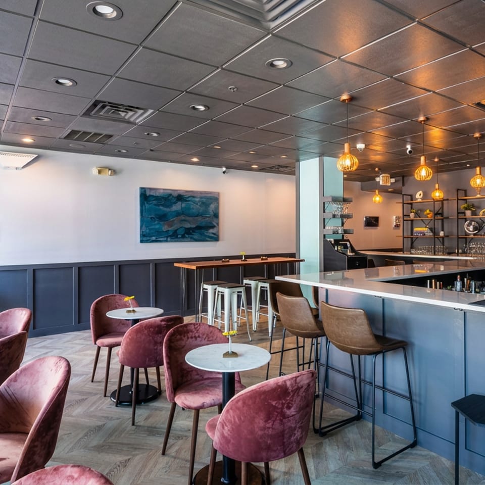 Bright contemporary restaurant interior with velvet chairs, marble-topped tables, a sleek bar, pendant lights, and blue wall artwork.