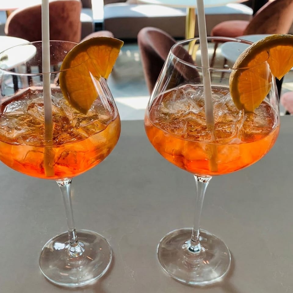 Two orange cocktails with ice, citrus garnishes, and straws on a table in a bright, modern restaurant.